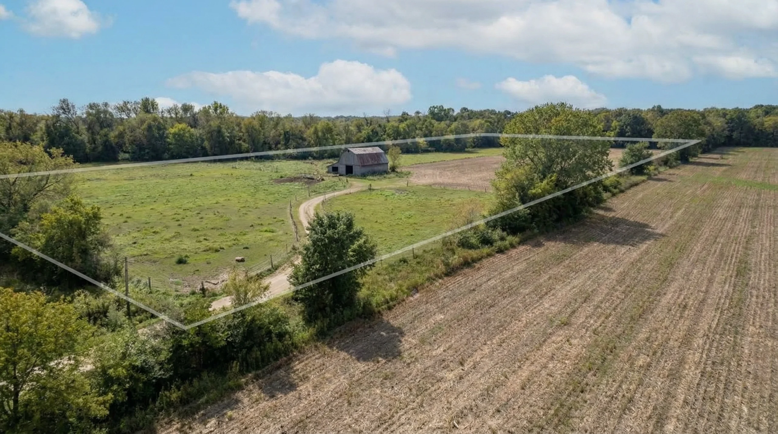 Drone view of a rural real estate property featuring wide open farmland, tree lines, and a small house along a quiet country road.