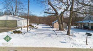 Snow-covered rural property with a country home, garage, and driveway surrounded by trees in a quiet countryside neighborhood